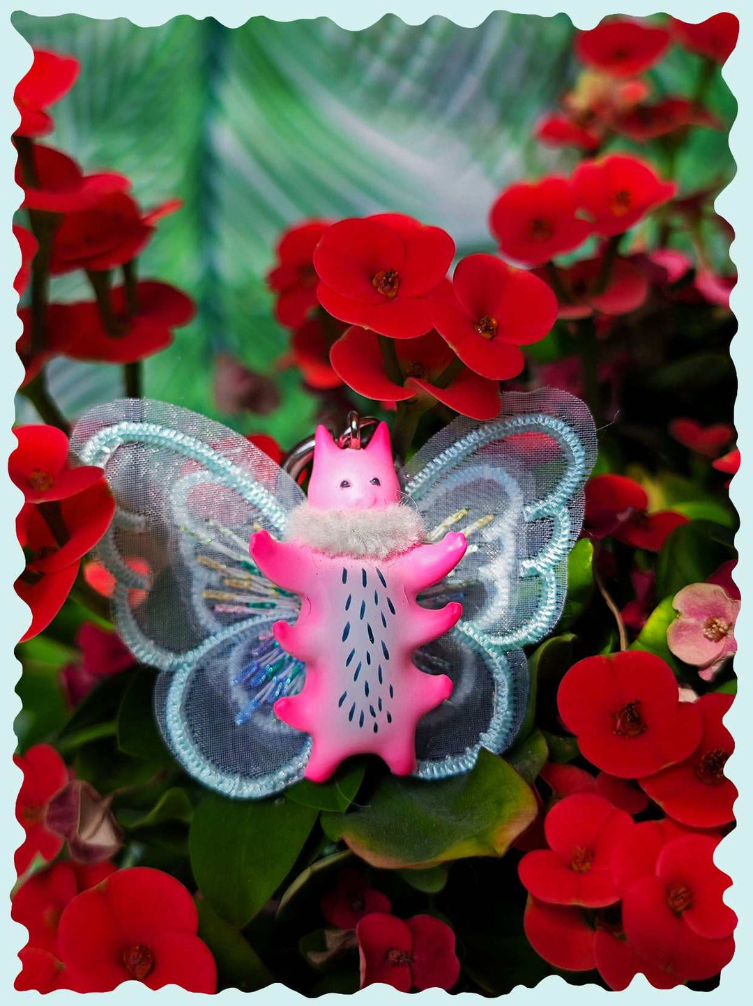 Butterfly cat with flamingo pink body and mint green wings amidst red flowers.