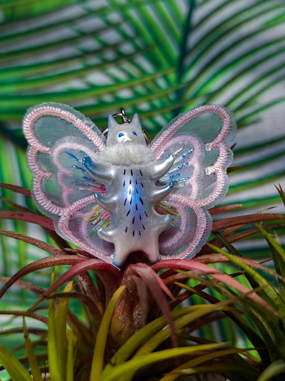 Butterfly cat with pearlescent wings and purple belly, wearing a fur snood necklace with earrings, surrounded by greenery.