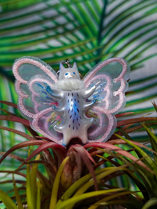 Butterfly cat with pearlescent wings and purple belly, wearing a fur snood necklace with earrings, surrounded by greenery.
